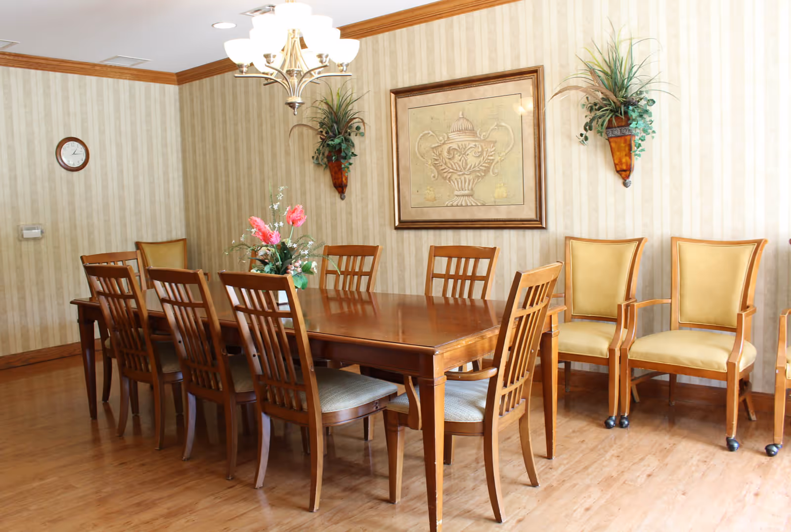 A dining room with a long wooden table surrounded by eight wooden chairs with cushioned seats. There are two additional cushioned armchairs with wheels against the wall. The room has striped wallpaper, a wooden floor, a chandelier hanging from the ceiling, a clock on the wall, two wall-mounted plant holders with greenery, and a framed artwork depicting a decorative urn.