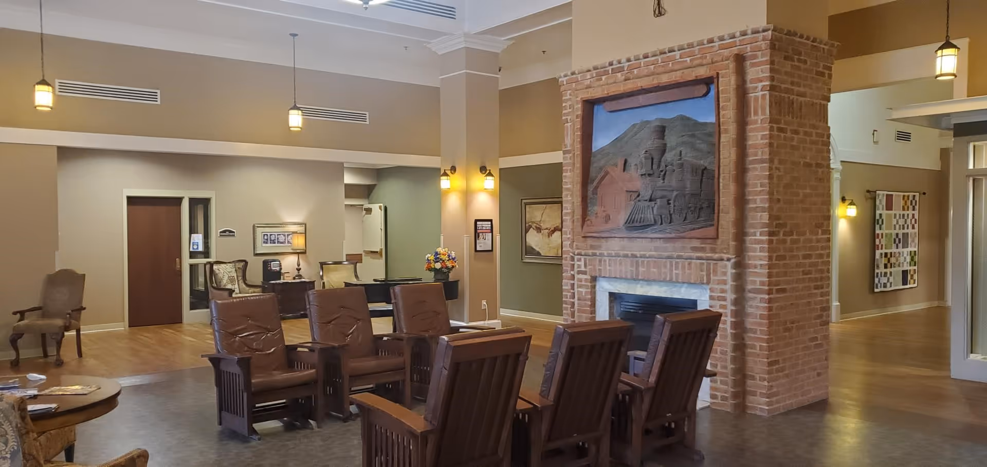 A spacious and well-lit common area in a retirement and assisted living facility featuring a brick fireplace with a large framed artwork above it. The room has several wooden chairs with cushions arranged around the fireplace, a round wooden table with magazines, and additional seating along the walls. The walls are painted in neutral tones with soft lighting from hanging and wall-mounted fixtures. The flooring is a combination of wood and tile, and there are decorative elements such as framed pictures and a flower arrangement on a side table.