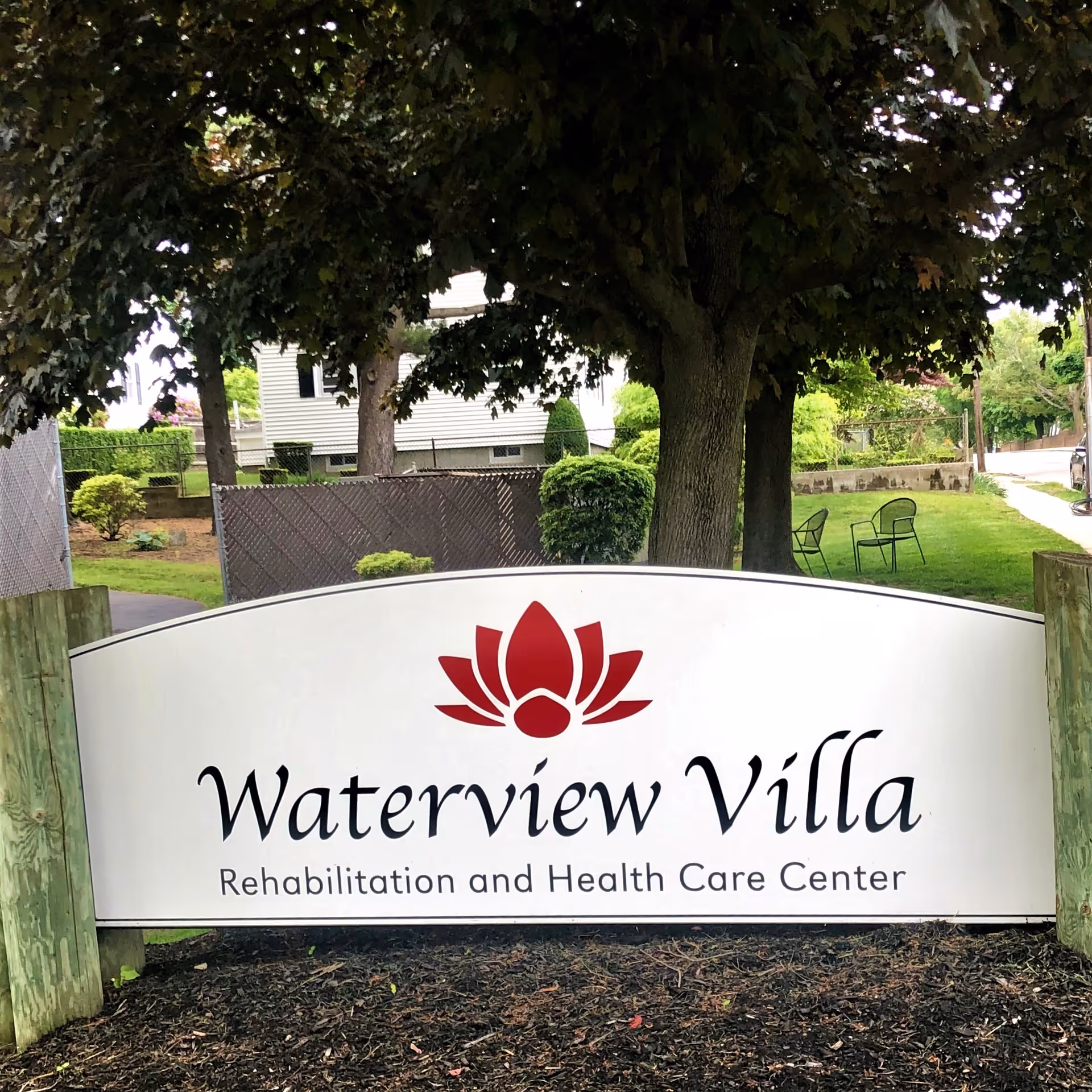 Outdoor sign for Waterview Villa Rehabilitation and Health Care Center with a red lotus logo, surrounded by trees, grass, and outdoor chairs in the background.