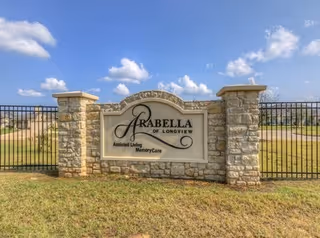 Stone sign for Arabella of Longview Assisted Living and Memory Care set in a grassy area with a black metal fence behind it under a blue sky with scattered clouds.