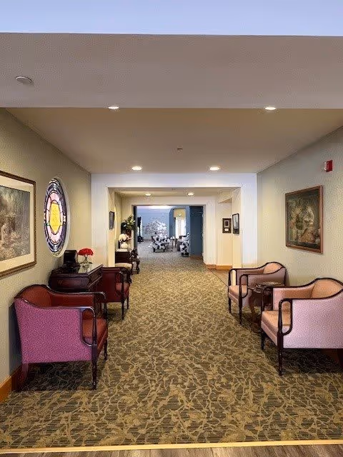A carpeted hallway in a senior living community with upholstered chairs and small tables along both walls. The walls are decorated with framed artwork and a circular stained glass window. The hallway leads to a well-lit common area with more seating visible in the background.