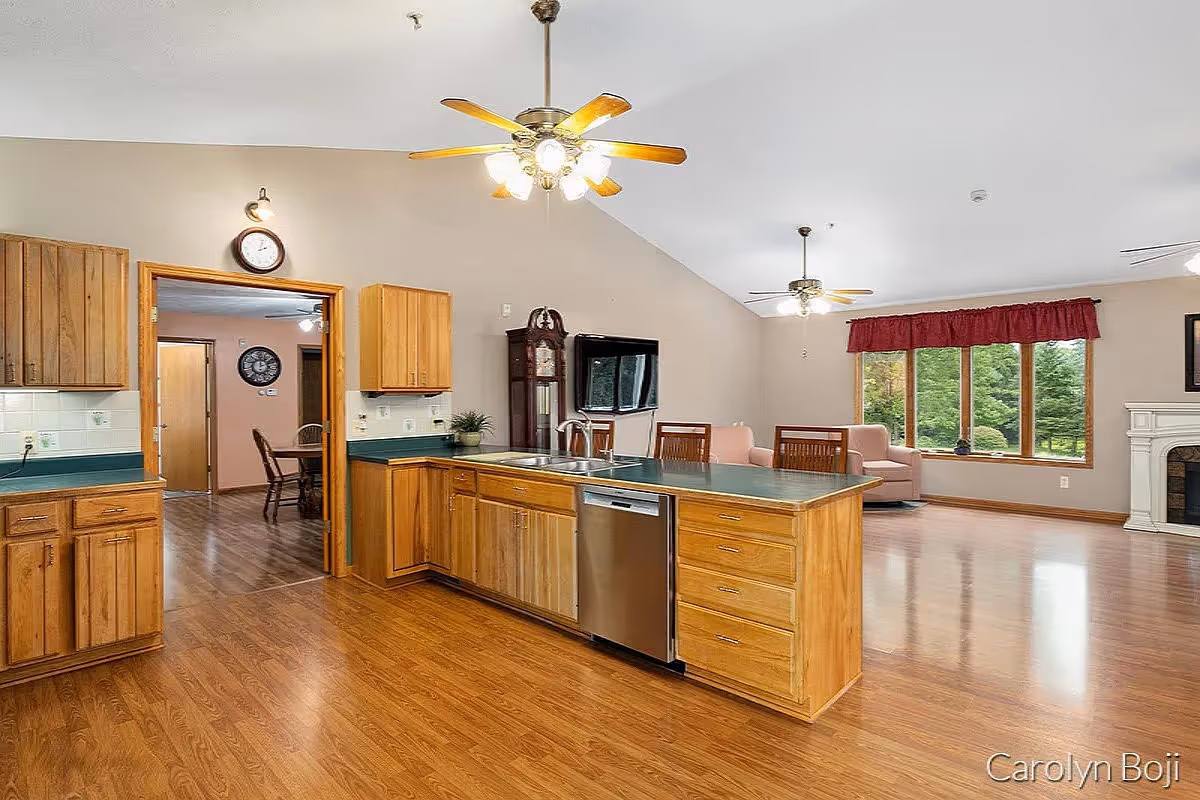 Spacious open-concept kitchen and living area with wooden floors and cabinetry. The kitchen features a large island with a sink and dishwasher, ceiling fans with lights, and a wall-mounted TV. The living area has large windows with red valances, a white fireplace, and light pink armchairs. A grandfather clock is positioned near the TV, and a dining area is visible through an open doorway.