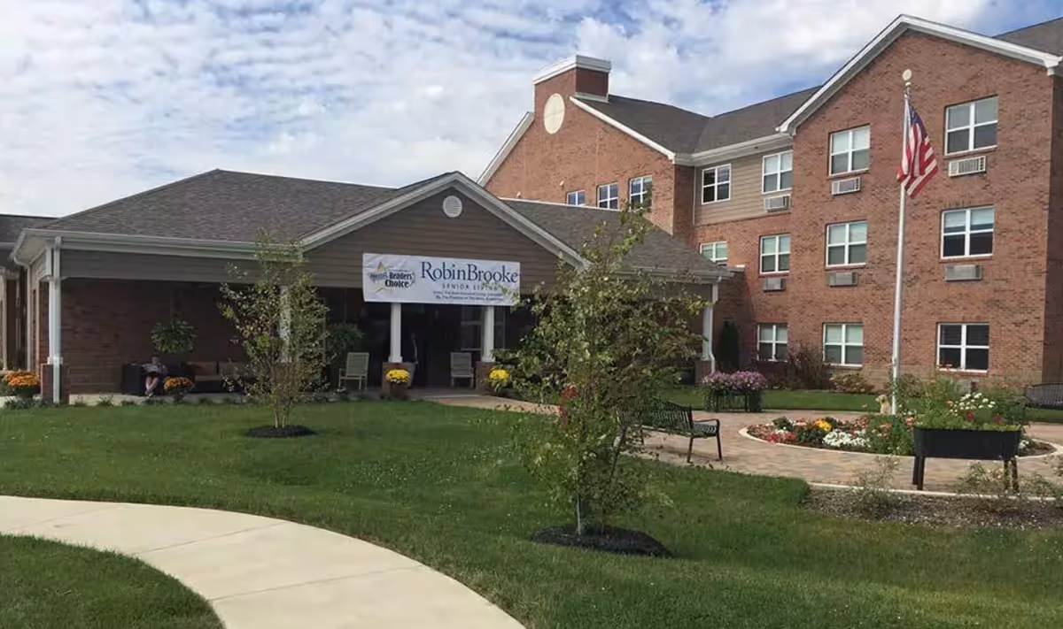 Exterior view of RobinBrooke Senior Living facility showing a brick building with multiple windows, a covered entrance with a sign, a flagpole with an American flag, green lawn, small trees, benches, and a circular flower bed.