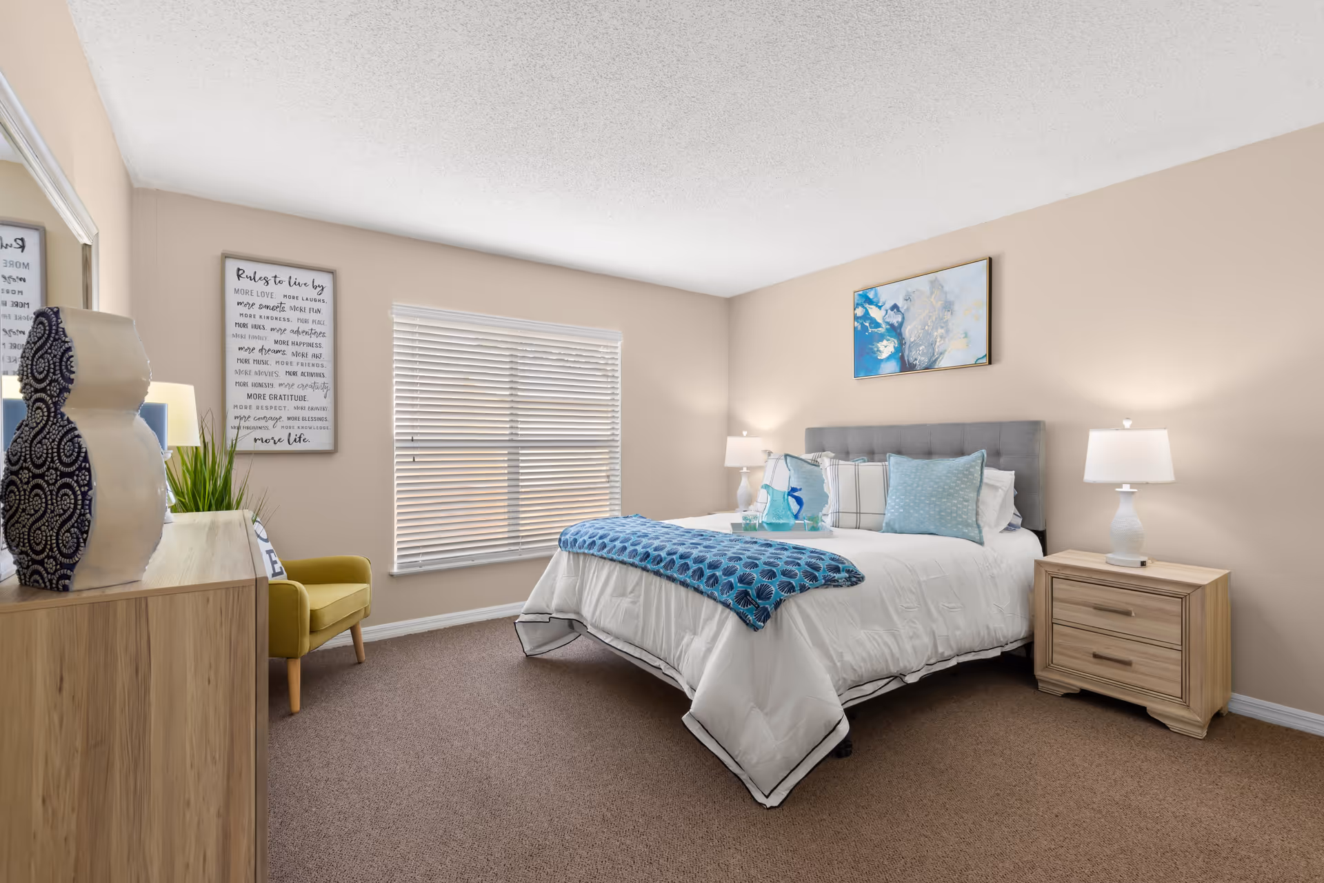 A modern bedroom featuring a queen-sized bed with a blue decorative throw, two bedside lamps, and a stylish dresser. The room has light-colored walls, a large window with blinds, and a piece of artwork above the bed.