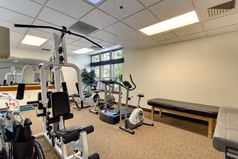 Small fitness room with weight machines, exercise bikes, a treadmill and benches against a mirrored wall and windows.
