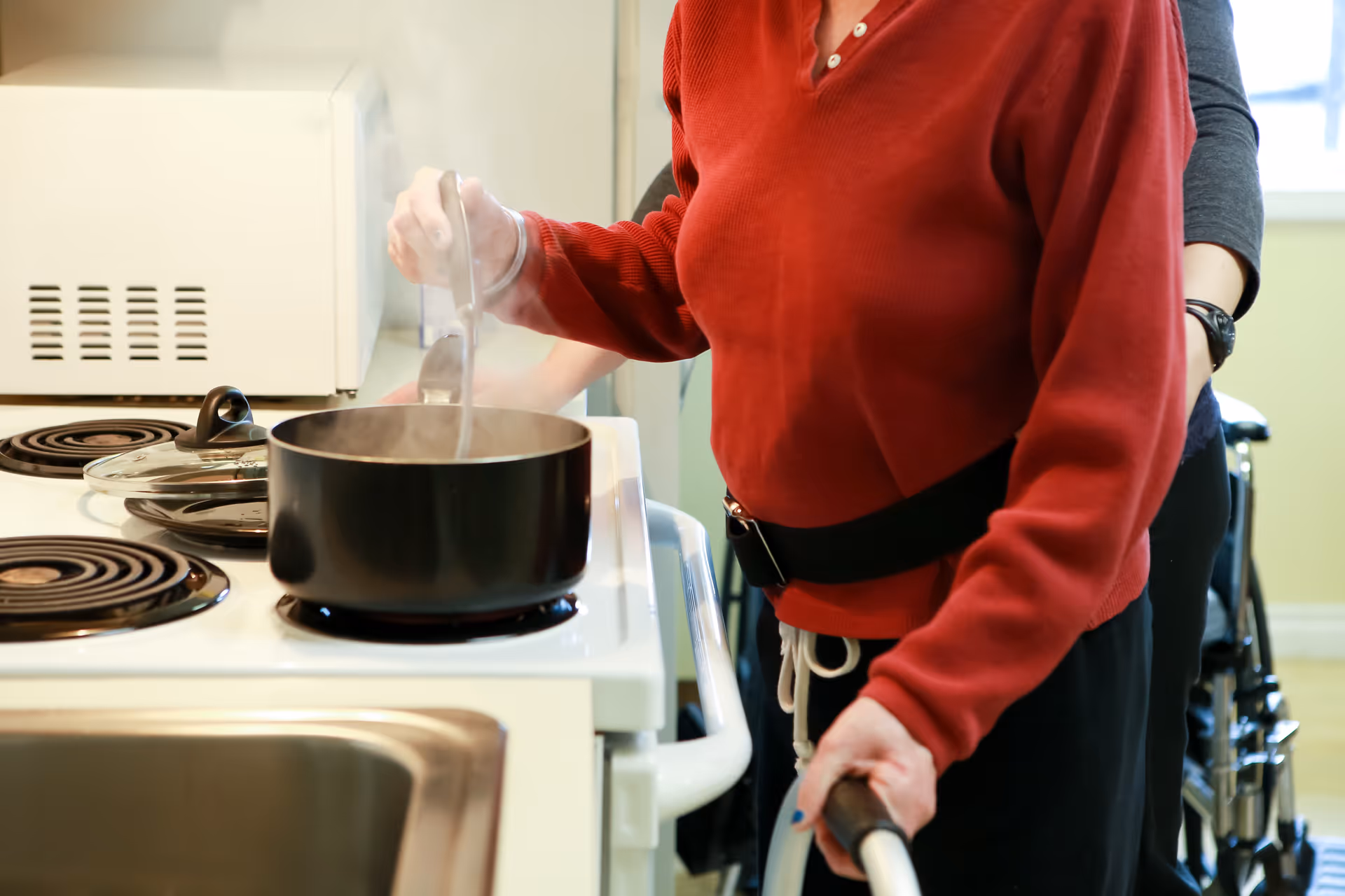 An elderly person wearing a red sweater is stirring a pot on a stove with assistance from another person standing behind them. The scene takes place in a kitchen with a microwave and stove visible.
