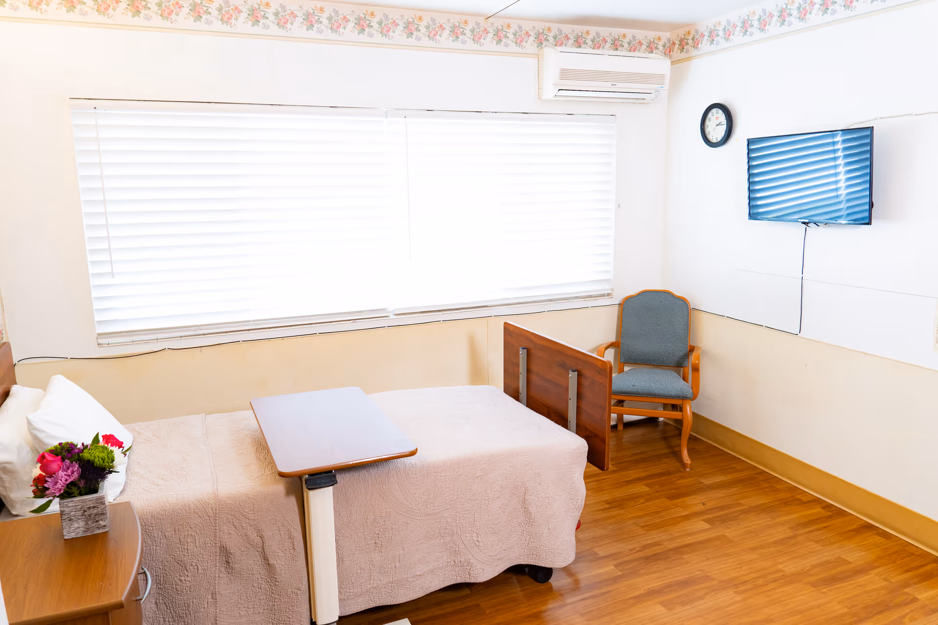 A simple, clean patient room with a single bed covered in a light beige quilt, a small overbed table, a wooden nightstand with a vase of flowers, a blue upholstered chair, a wall-mounted flat-screen TV, a clock, and a large window with white blinds. The room has wood flooring and floral wallpaper border near the ceiling.