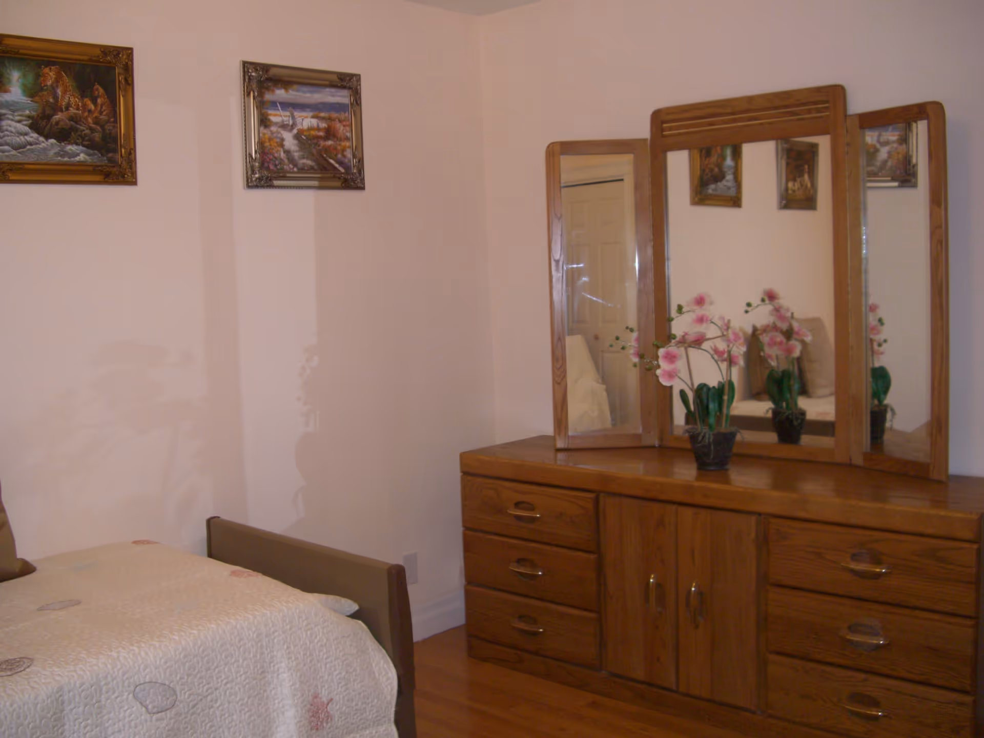 A bedroom corner featuring a wooden dresser with a three-panel mirror and a potted pink orchid on top. To the left, part of a bed with a light-colored quilt is visible. Two framed paintings hang on the light-colored wall above the bed.