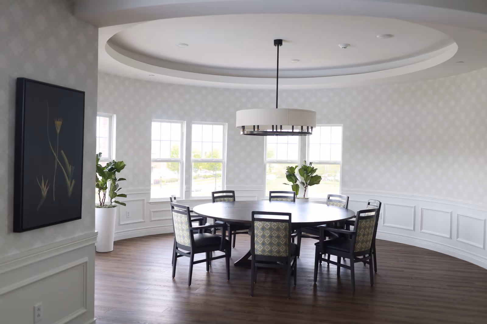 A bright, circular dining room with a large round wooden table surrounded by eight chairs with patterned backs. The room features large windows letting in natural light, white wainscoting on the walls, a modern circular ceiling light fixture, and two potted plants near the windows. A framed artwork is visible on the left wall.