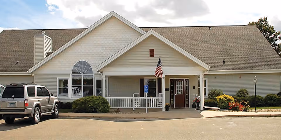 Front exterior view of a single-story senior living facility building with a peaked roof, a covered entrance with white railings, an American flag, and a parked SUV in front. There are bushes and flowers along the building's facade under a partly cloudy sky.