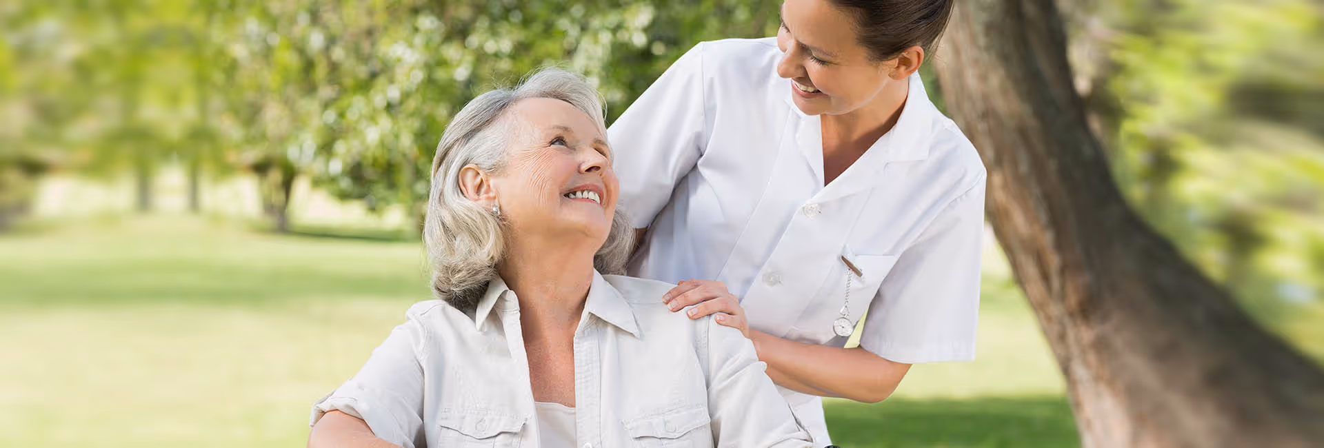 A smiling elderly woman sitting outdoors in a wheelchair, being gently pushed by a smiling female caregiver dressed in white uniform, with green trees and grass in the background.