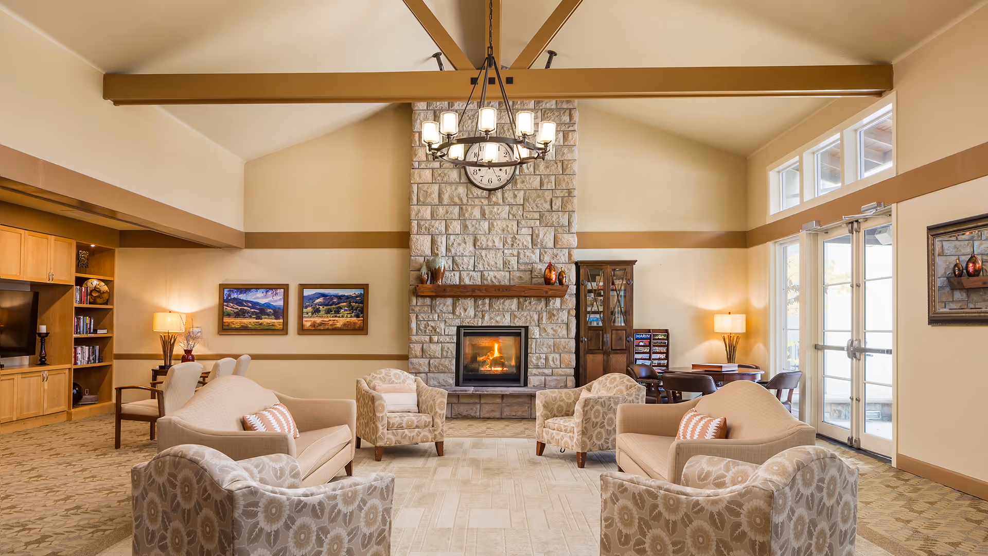 A spacious and well-lit living room area with a high vaulted ceiling and exposed wooden beams. The room features a stone fireplace with a clock above it, surrounded by two armchairs and two sofas with patterned cushions. There are large windows and glass doors on the right side letting in natural light. On the left side, there is a built-in wooden entertainment center with a TV and shelves. The room is decorated with framed landscape paintings, lamps, and a small table with chairs near the windows.