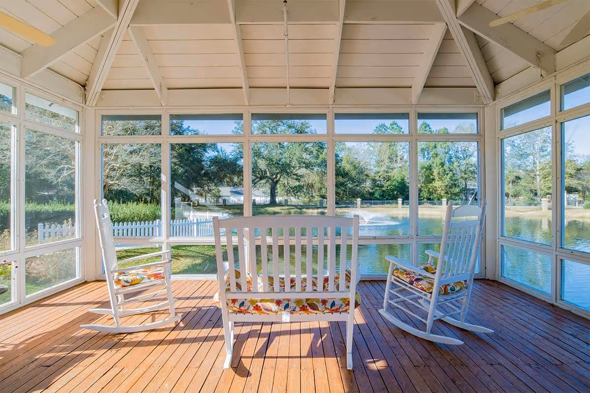 A bright screened-in porch with wooden flooring and a vaulted ceiling, furnished with two white rocking chairs and a white bench, all with colorful floral cushions. The porch overlooks a pond with a fountain and is surrounded by trees and a white picket fence.