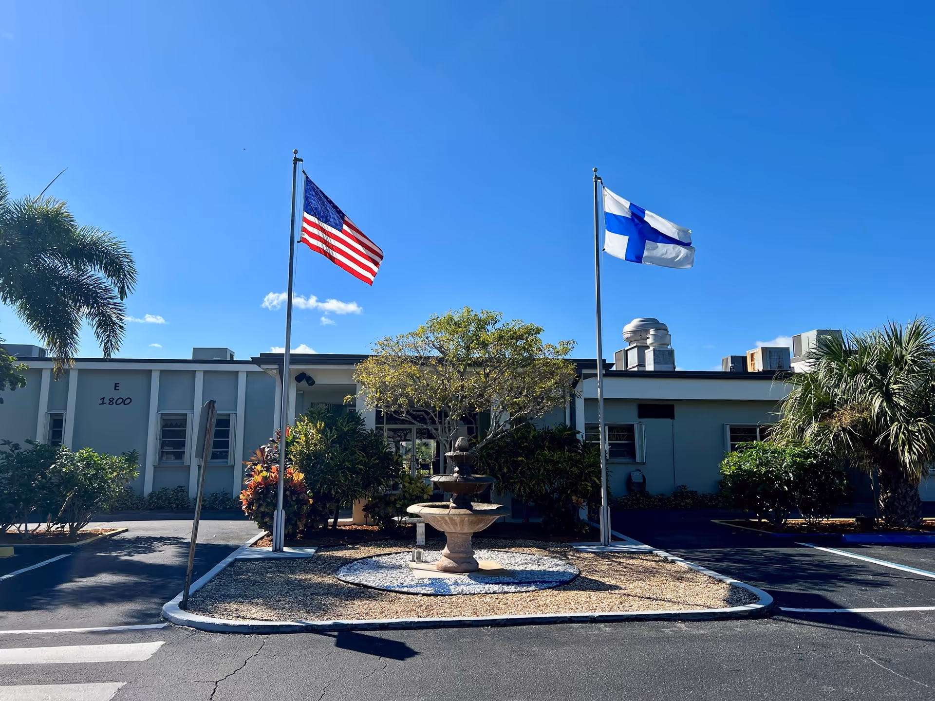 Exterior view of a single-story building with two flagpoles in front, one displaying the American flag and the other displaying the Finnish flag. There is a small fountain surrounded by landscaping with bushes and trees in front of the building. The sky is clear and blue.