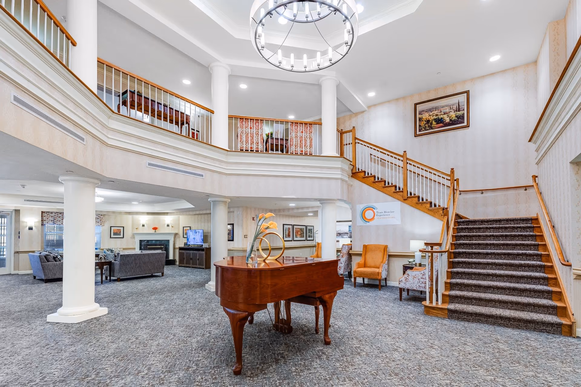 Spacious senior living facility common area with a grand piano in the center, carpeted floors, a staircase with wooden handrails leading to an upper balcony, comfortable seating areas with armchairs and sofas, a fireplace, and framed artwork on the walls under bright ceiling lights.