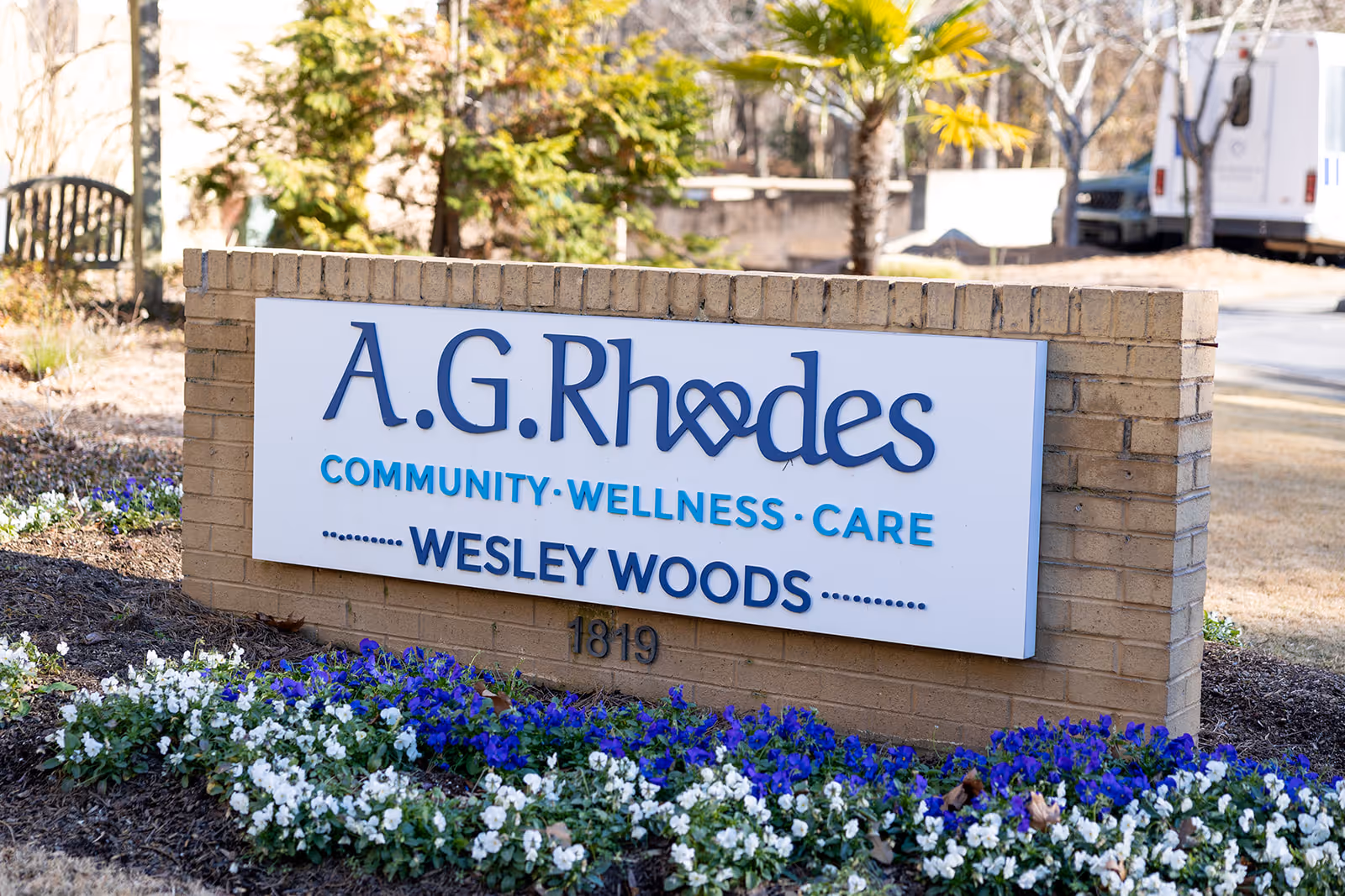 Brick entrance sign for A.G. Rhodes Wesley Woods with blue and white flowers planted at its base.