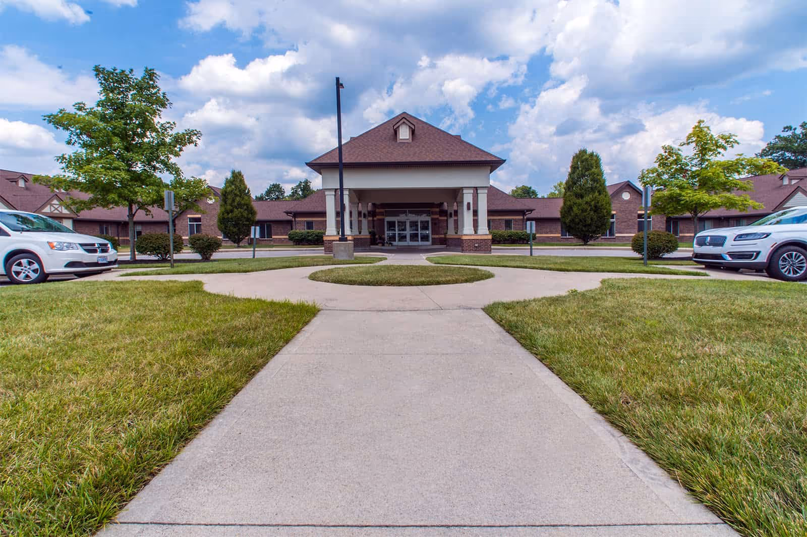 Front exterior view of Forest Hills Healthcare Center showing a covered entrance with a peaked roof, surrounded by a circular driveway, green lawns, trees, and parked cars under a partly cloudy sky.