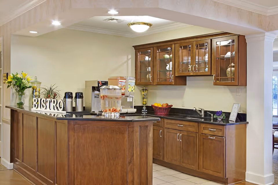 A small kitchen area with wooden cabinets and a black countertop. On the counter, there are beverage dispensers, a water container with fruit slices, a basket of fruit, and decorative items including a vase with flowers and a sign that reads 'BISTRO'. The space is well-lit with ceiling lights and has a clean, welcoming appearance.