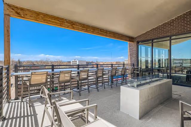 Outdoor balcony area with multiple chairs and a long counter facing a railing overlooking a cityscape. There is a modern rectangular fire pit in the center and glass doors leading to an interior space.