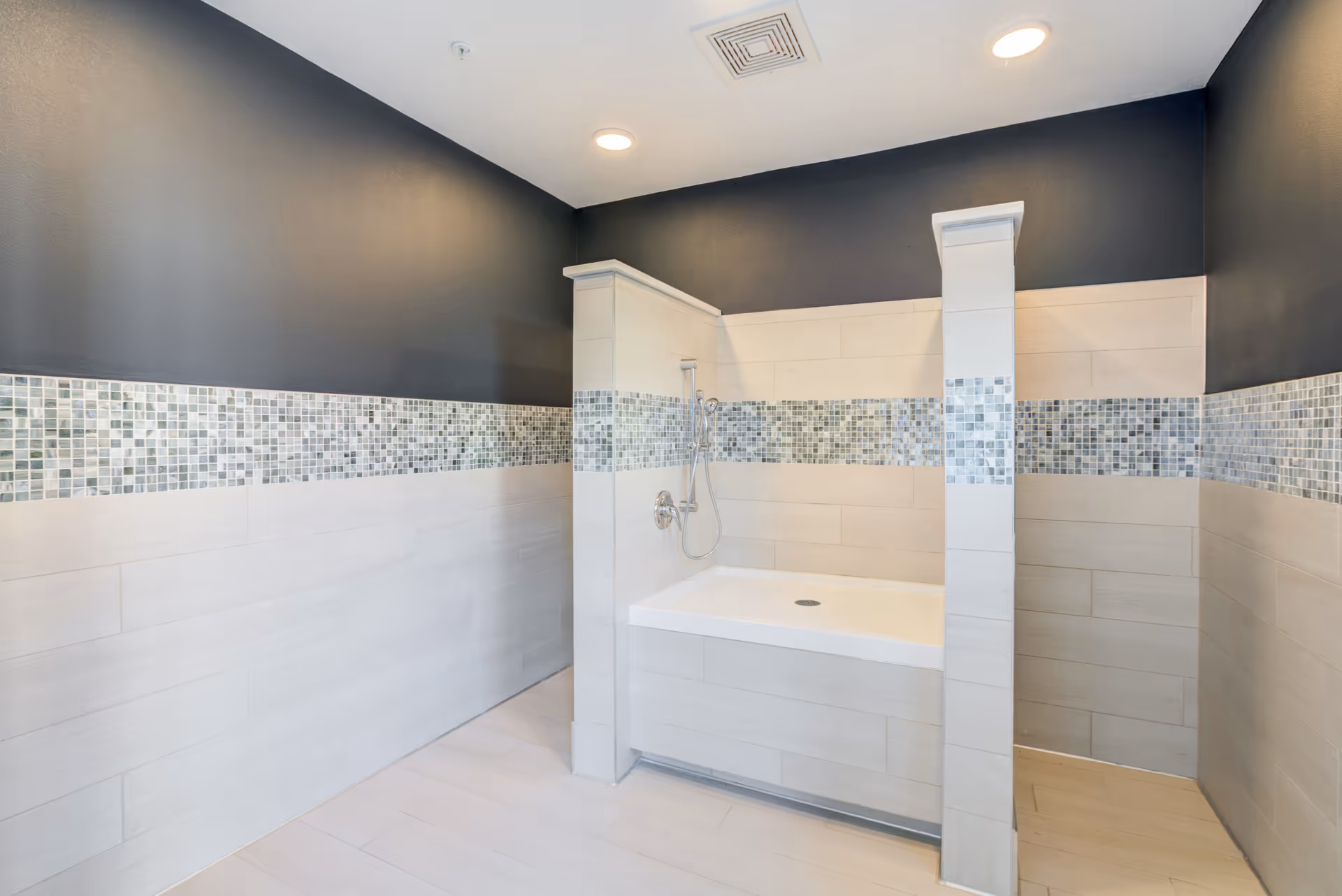 A modern bathtub/shower alcove with light tile, a horizontal mosaic tile band, and dark upper walls under recessed lighting.