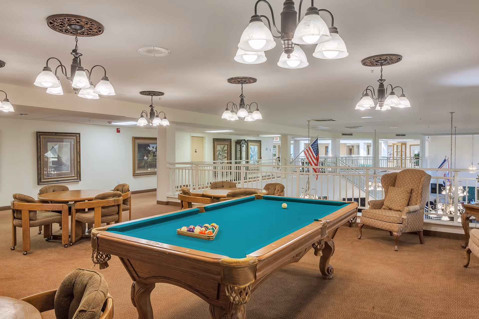 A spacious indoor recreational area with a pool table in the center, surrounded by cushioned chairs and tables. The room is well-lit with multiple ceiling light fixtures, and there are framed paintings on the walls. An American flag is visible in the background near a railing overlooking a lower level.