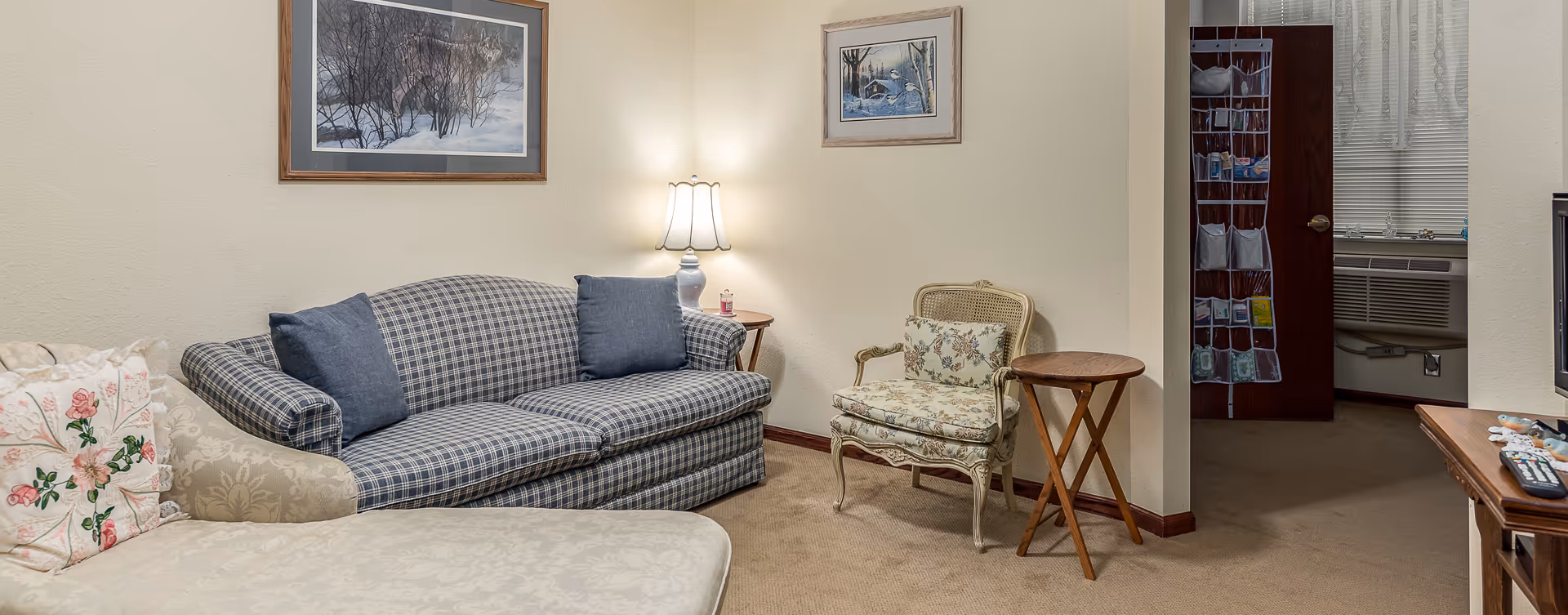 A cozy living room area with a blue plaid sofa with two blue cushions, a floral upholstered armchair with a matching cushion, a beige chaise lounge with a floral pillow, a wooden side table, and a lit table lamp on a small round table. Two framed pictures hang on the cream-colored walls. A doorway leads to another room with a window air conditioner and a door organizer hanging on the door.