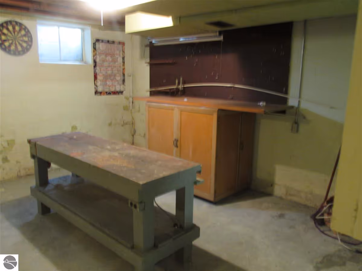 Dim basement room with a central wooden workbench, a wall cabinet, a small window, and a dartboard on the wall.