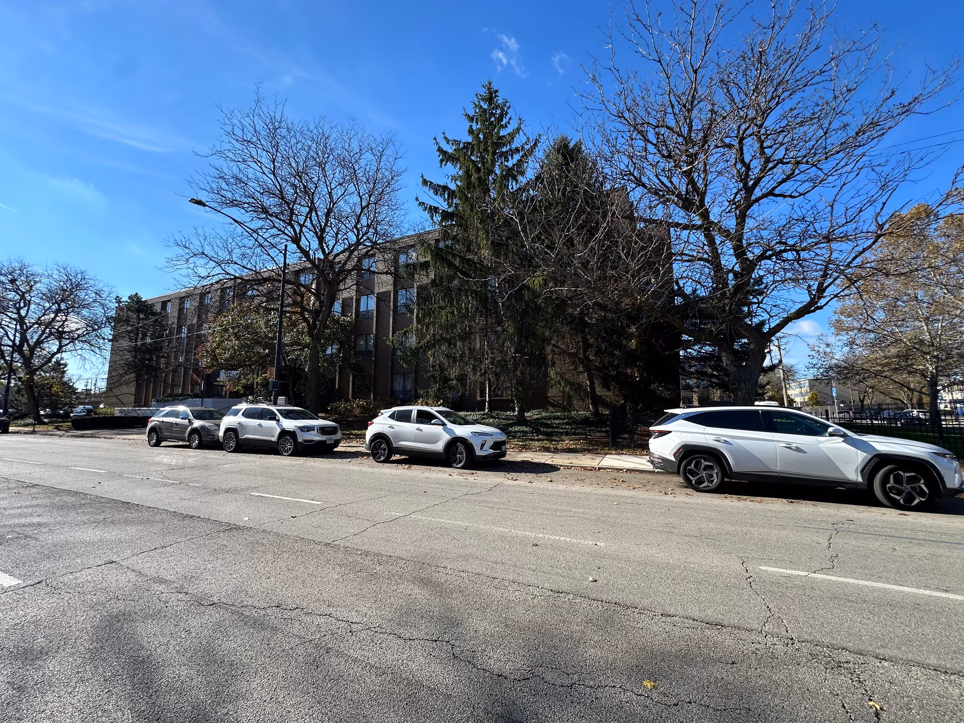 Street view of a multi-story brick building with parked cars and leafless trees under a clear blue sky.