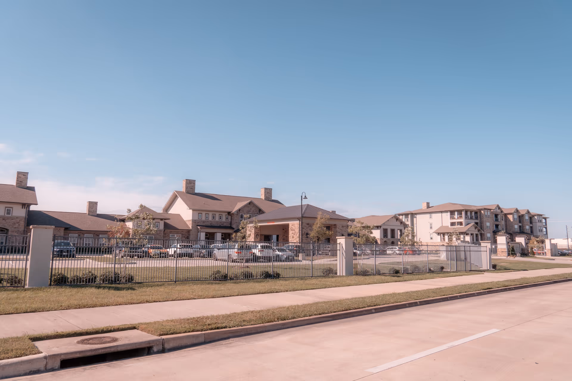 Exterior view of The Lodge at Pine Creek facility showing multiple connected buildings with stone and beige siding, a parking lot with several cars, a metal fence, and a clear blue sky.