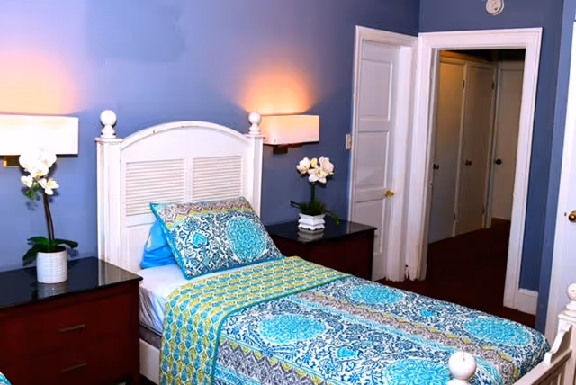 A neatly made single bed with a white headboard and colorful blue, green, and gray patterned bedding in a room with blue walls. There are two dark wooden nightstands on either side of the bed, each with a white orchid plant and a wall-mounted lamp above. An open doorway leads to a hallway with white doors.