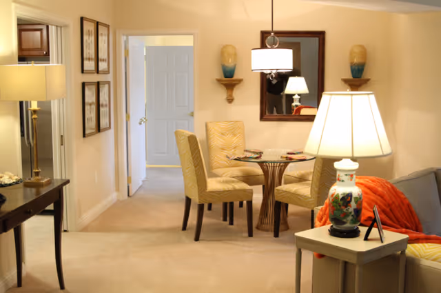 A cozy interior living space featuring a small round glass dining table with four beige upholstered chairs. A large mirror hangs on the wall above the table, flanked by two decorative wall shelves with vases. To the right, a side table with a decorative lamp and an orange throw blanket on a gray sofa are visible. The room is softly lit by multiple lamps, creating a warm and inviting atmosphere. A hallway with an open door leads to another room in the background.