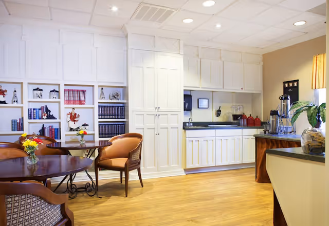 Bright dining/common area with round tables and chairs, built-in white cabinetry, bookshelves, and a small kitchenette.