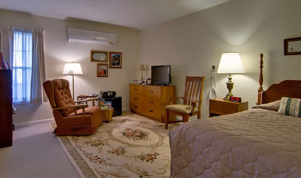 A cozy senior living bedroom featuring a bed with a quilted bedspread and a decorative pillow, a wooden nightstand with a lamp and digital clock, a wooden dresser with a small TV on top, a wooden chair, and a comfortable upholstered armchair next to a floor lamp. The room has a window with curtains, framed pictures on the wall, and a patterned area rug covering the carpeted floor.