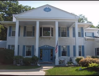 Front exterior of a two-story white manor-style building with tall columns, blue shutters, and an American flag by the entrance.