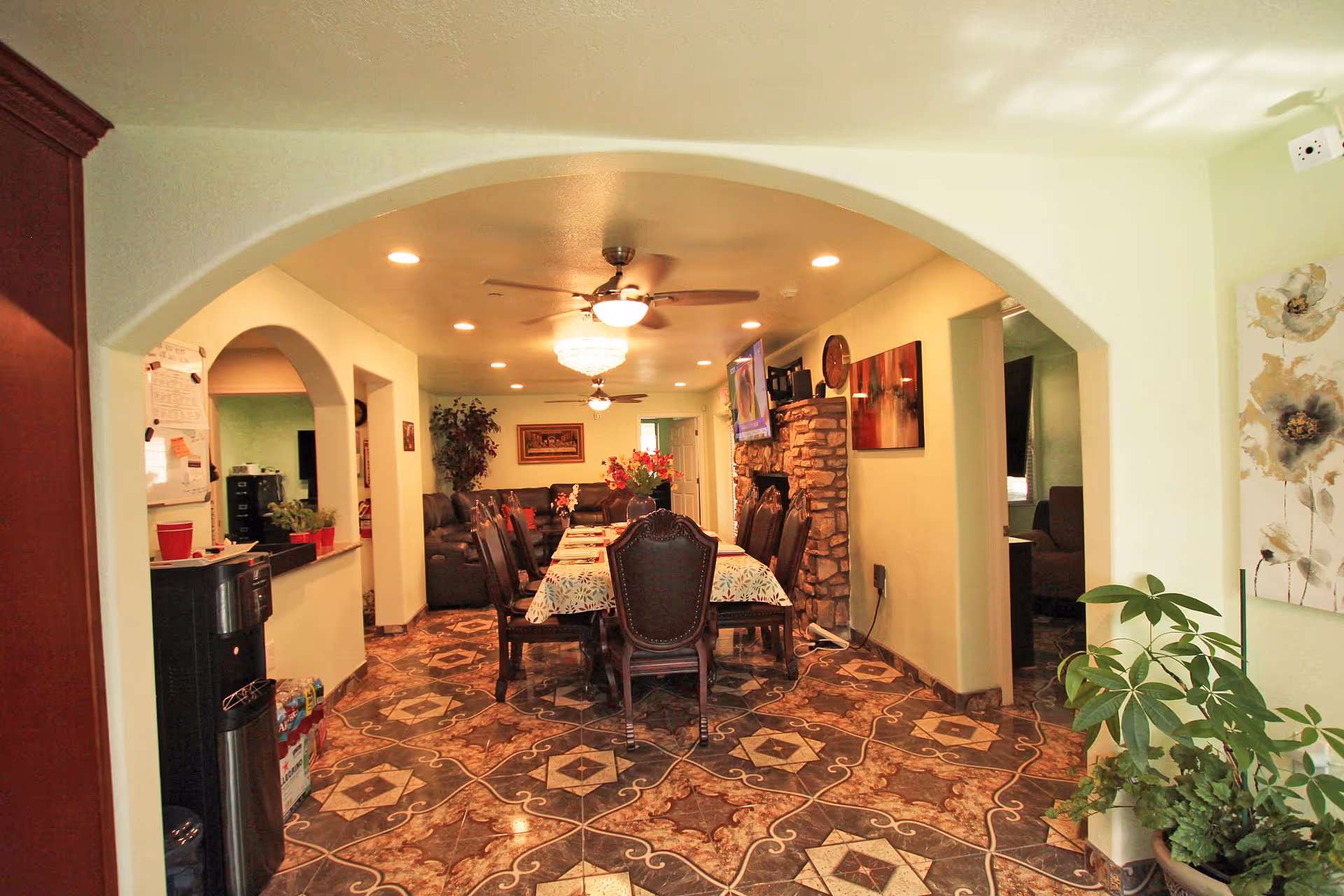 Interior view of a senior living facility dining and living area with a long dining table covered with a floral tablecloth and surrounded by wooden chairs. The room features patterned tile flooring, a stone fireplace with a mounted TV above it, ceiling fans, recessed lighting, and decorative plants and artwork on the walls.
