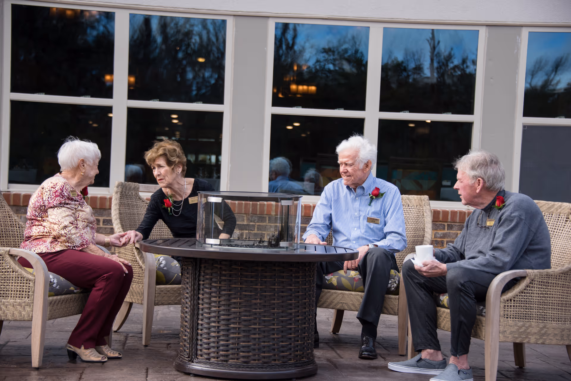 Four elderly residents seated around a round outdoor patio table, conversing beside the building's windows.