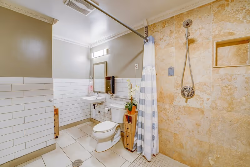A clean and well-lit bathroom featuring a toilet, a wall-mounted sink with a mirror above it, a shower area with beige tiled walls and a striped shower curtain, and a small wooden storage unit with a potted plant on top.