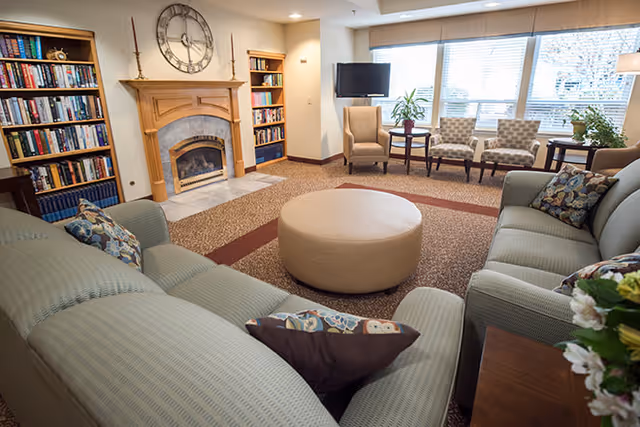 A cozy living room area with two green sofas facing each other, a round beige ottoman in the center, a fireplace with a large clock above it, bookshelves filled with books, and a seating area with three chairs near large windows letting in natural light.