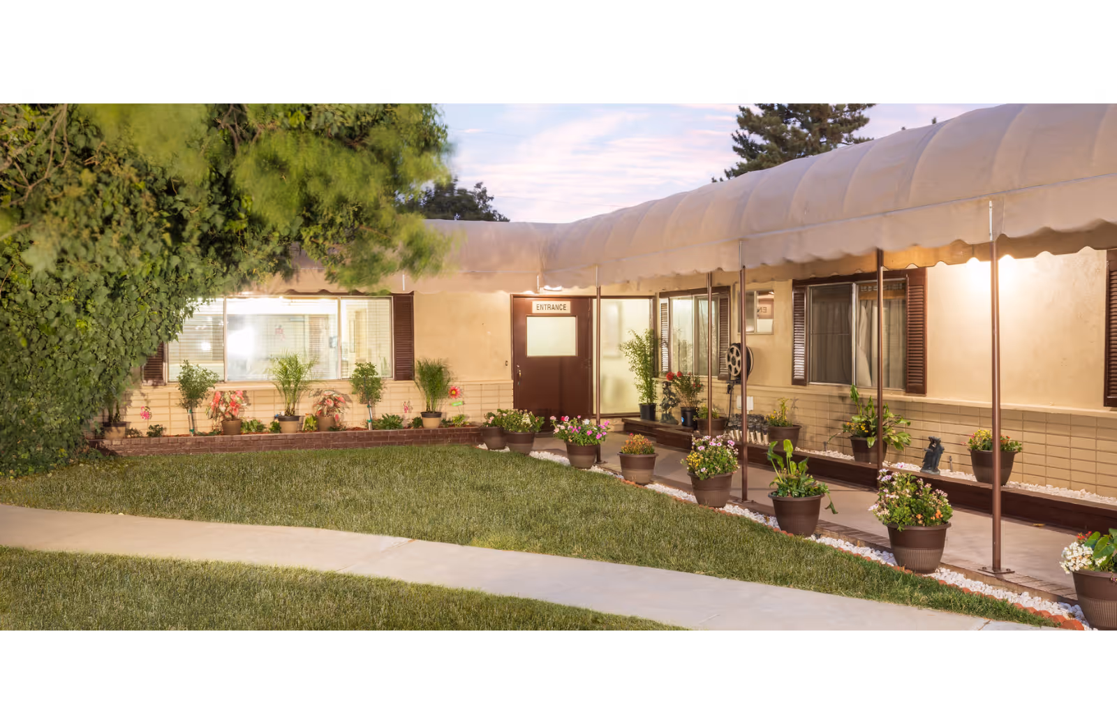 Covered entrance and walkway of a single-story facility with potted plants, lawn, and windows.