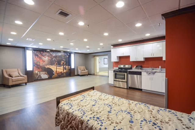 A spacious room in a senior living facility featuring a bed with a patterned bedspread in the foreground, a kitchenette with white cabinets, stainless steel appliances, and a red backsplash on the right side, and a seating area with two armchairs and a large wall mural depicting a scenic bridge and autumn trees in the background.