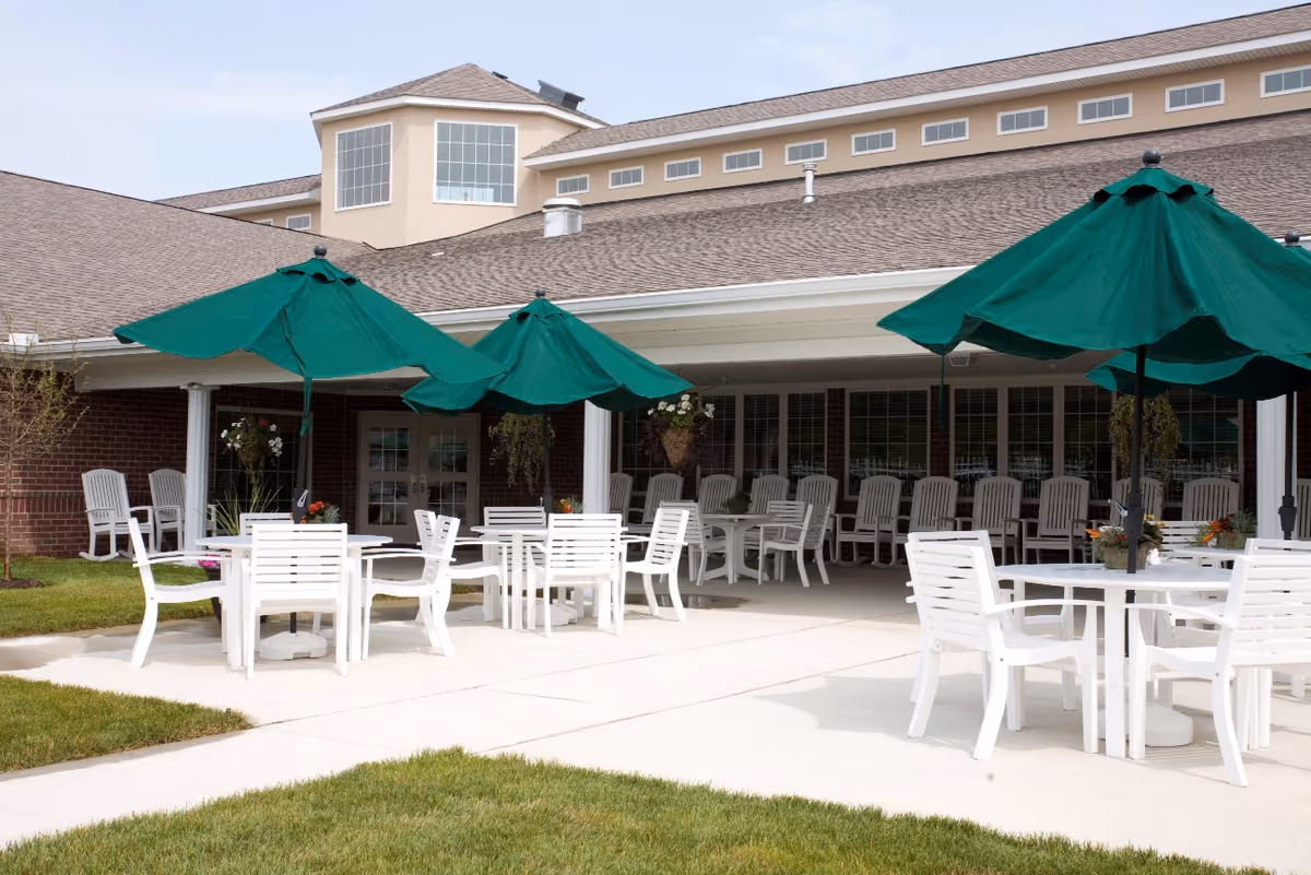 Outdoor patio area at Bridgewater Healthcare Center with white tables and chairs, green umbrellas providing shade, and a building with large windows and a covered porch in the background.