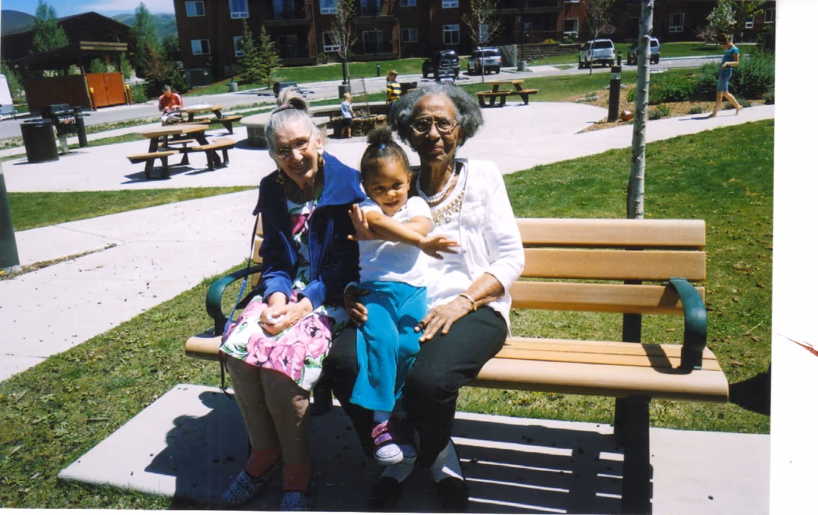 Two elderly women sitting on a park bench outdoors with a young child sitting on one woman's lap. In the background, there are picnic tables, a few people walking, and residential buildings.
