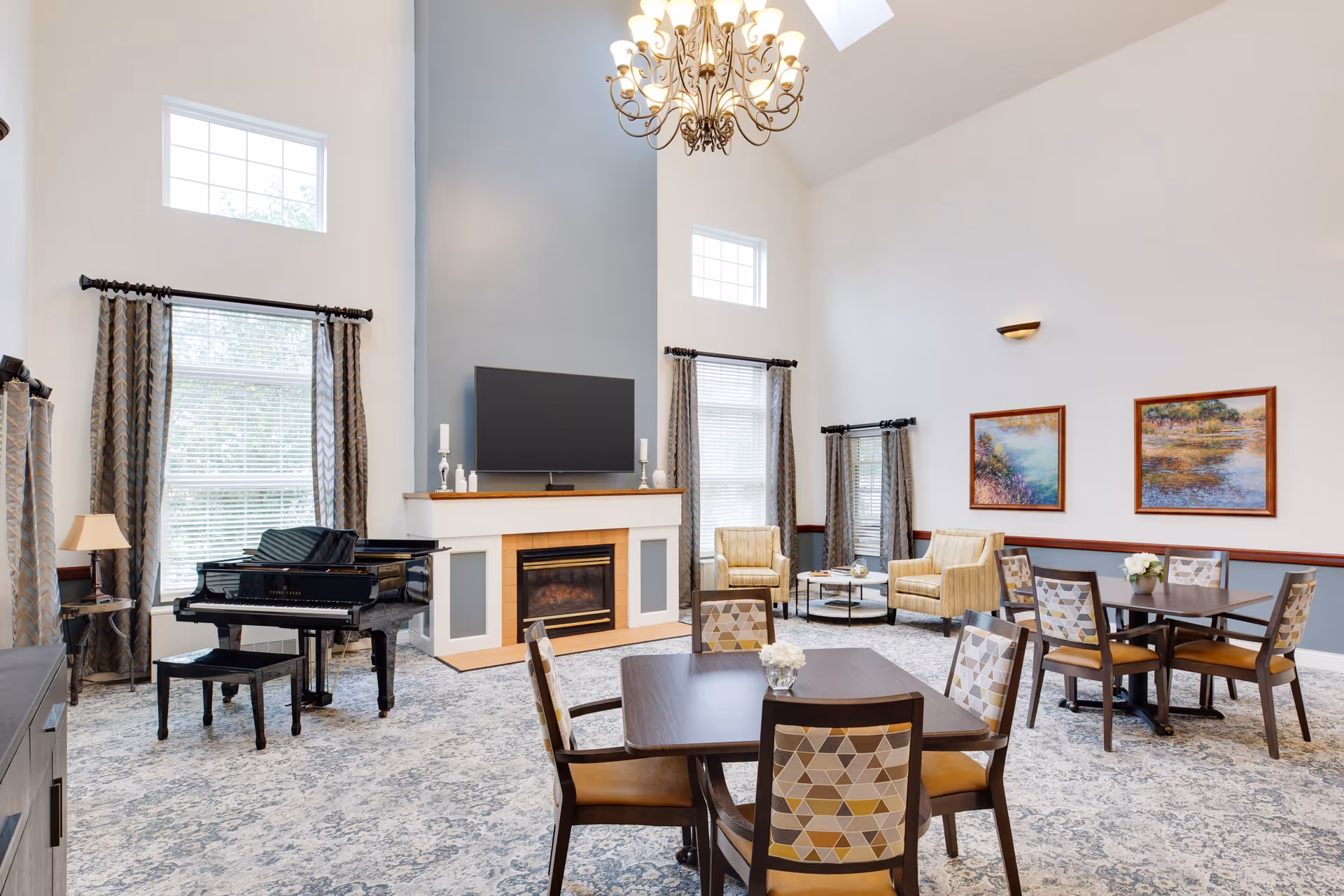 Spacious senior living common room with a grand piano, fireplace and TV, seating areas, and dining tables under a chandelier.