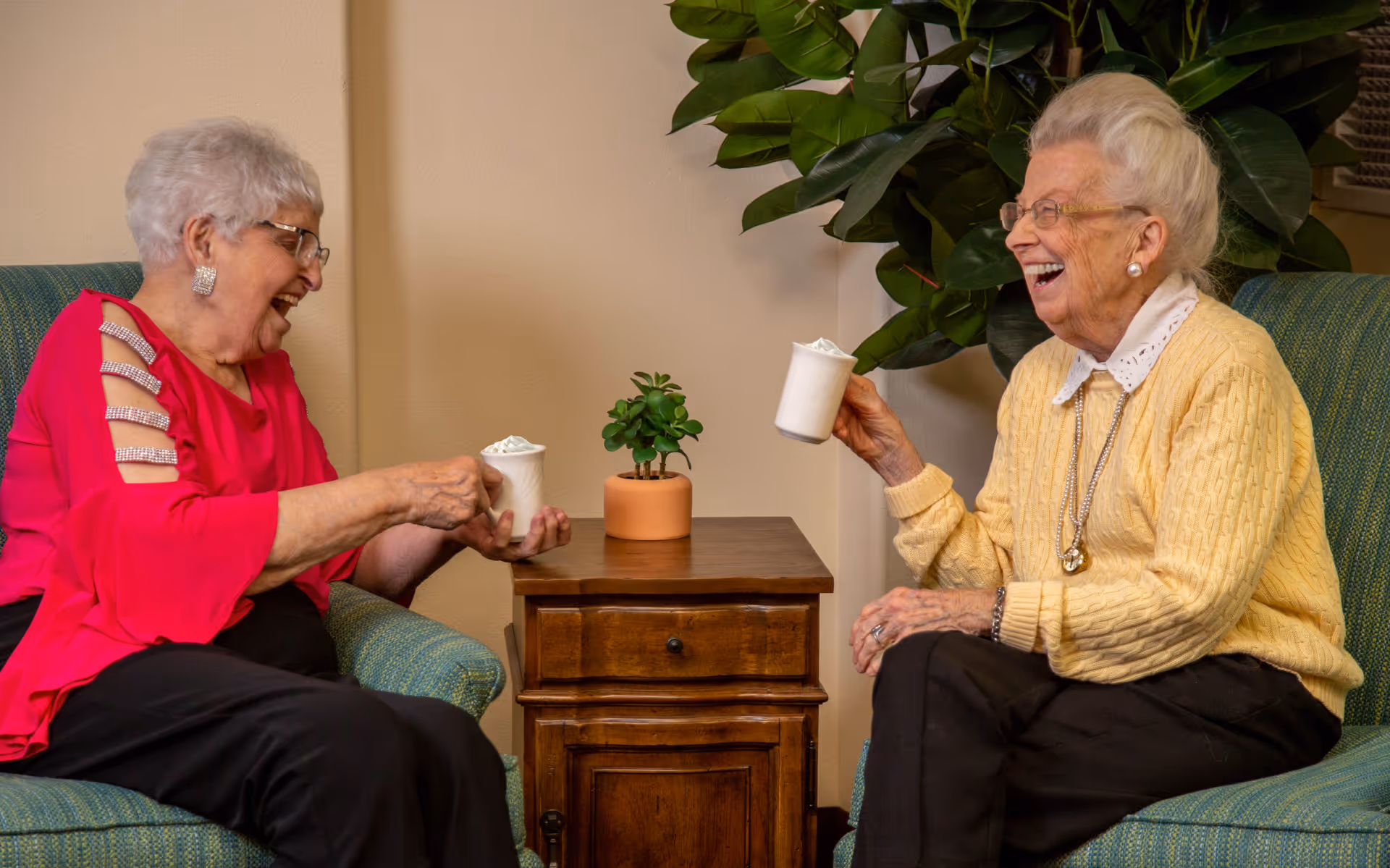 Two elderly women sitting in armchairs facing each other, laughing and holding white mugs with whipped cream. They are seated on either side of a small wooden table with a potted plant on it, in a cozy indoor setting with a large green plant in the background.
