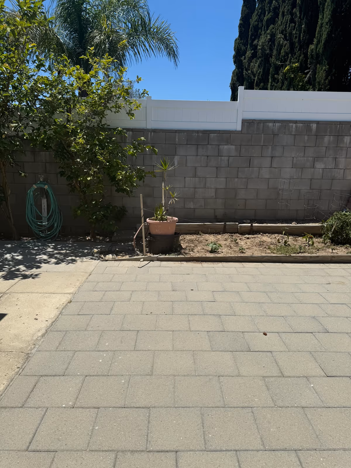 Outdoor patio area with a paved stone floor, a small garden bed with some plants, a potted plant, a garden hose coiled on a wall-mounted holder, and a concrete block wall topped with a white fence. Trees and a clear blue sky are visible in the background.