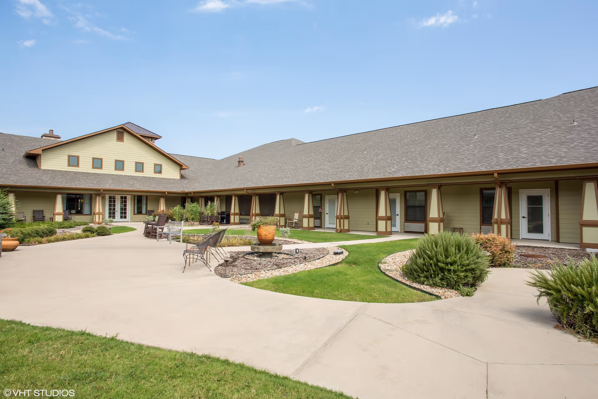 Courtyard of a single-story assisted living building with a paved walkway, seating, planters, and doors under a covered porch.