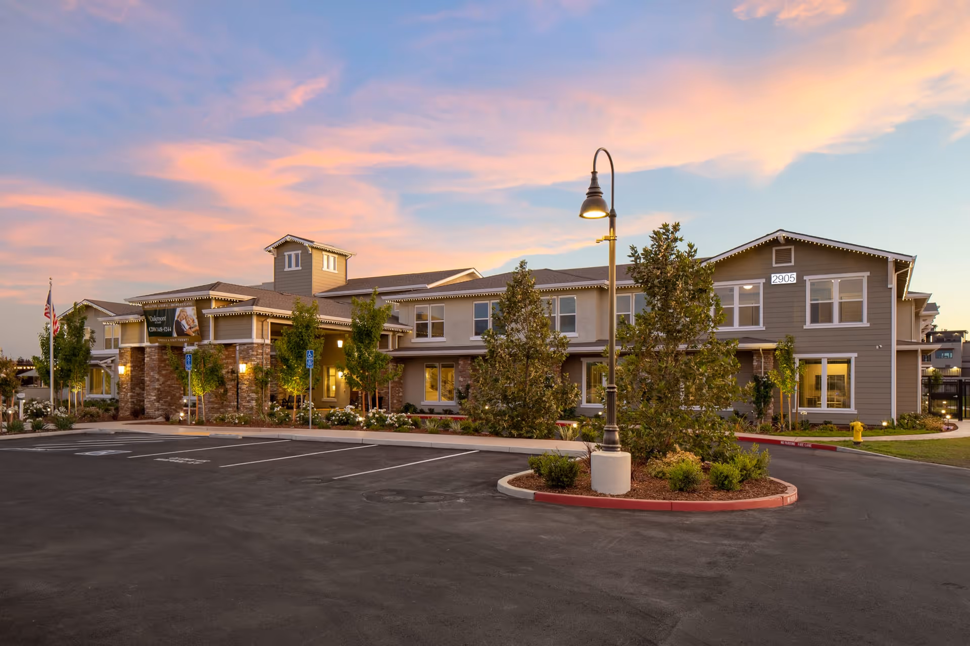 Exterior view of Oakmont of Lodi senior living facility at sunset, showing a two-story building with a stone and siding facade, landscaped greenery, a parking lot with handicap spaces, and a street lamp in the foreground.