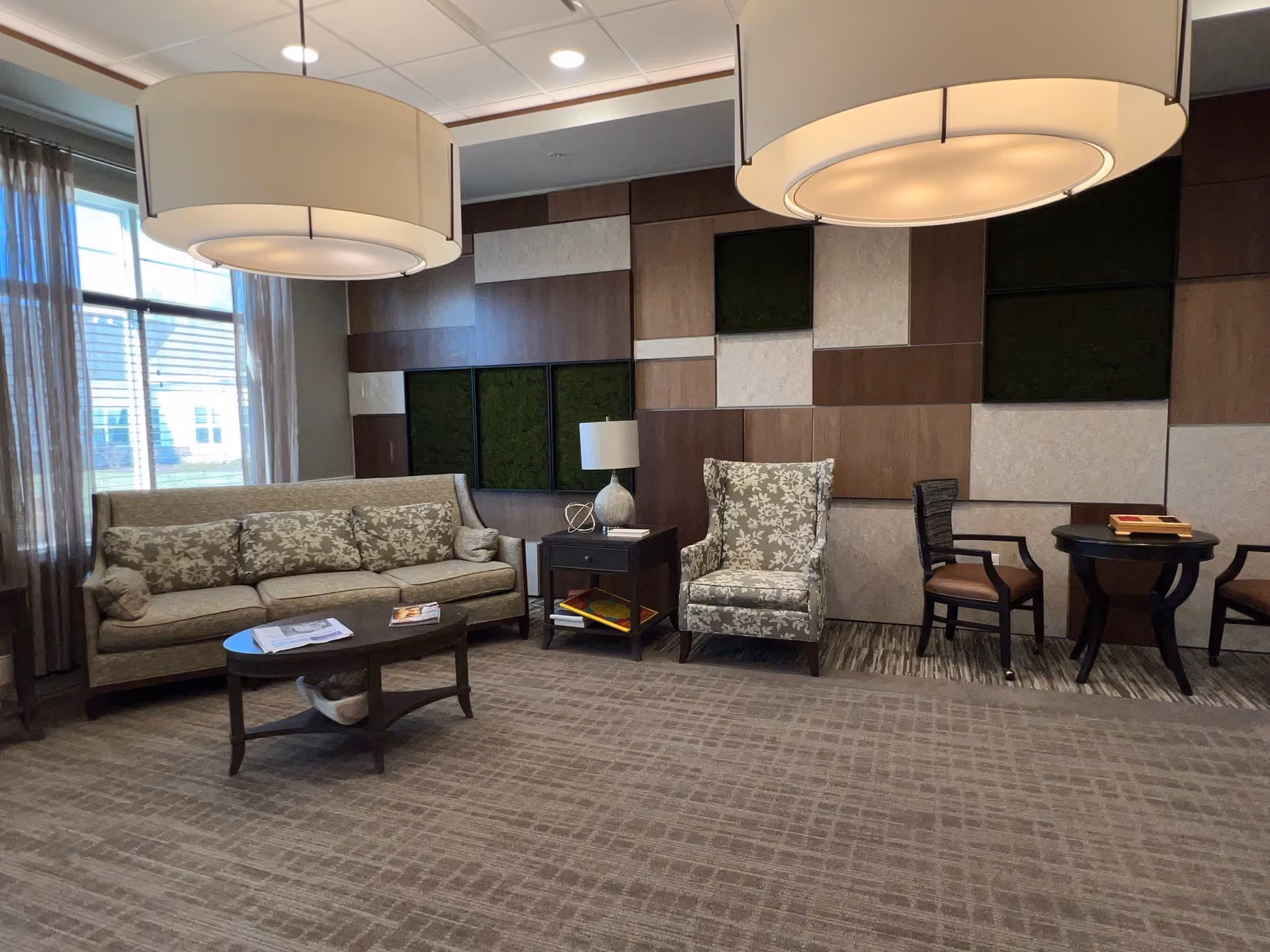 A cozy living room area with a floral-patterned sofa and matching armchair, a dark wooden coffee table with magazines, a side table with a lamp, and two wooden chairs with a small round table. The wall behind features a decorative panel design with wood and fabric squares. Large windows with sheer curtains allow natural light to fill the room. Two large round ceiling lights hang above.