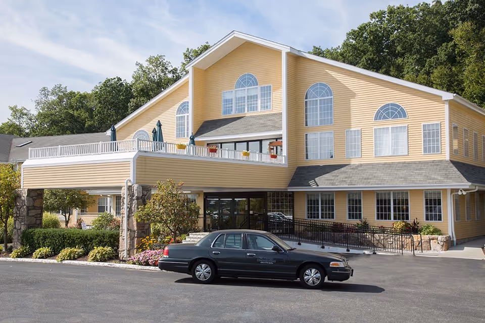 Yellow multi-story senior living building with arched windows, a covered entrance and a car parked in front.