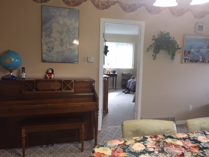 Interior view of a senior living facility room featuring a wooden piano with a globe and a small stuffed monkey on top. A floral garland hangs above the doorway leading to another room with a window and a person sitting on a chair. A table with a floral tablecloth and cushioned chairs are partially visible in the foreground. The walls are decorated with framed pictures and a hanging plant.